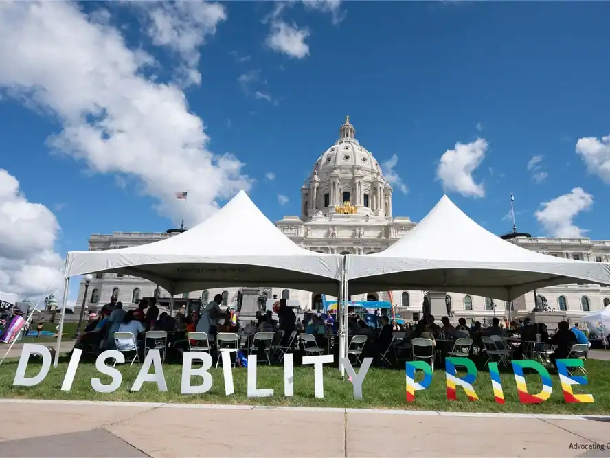 A tent with people and Disabaility Pride in large letters in from of a capitol building.